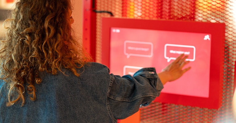 Effective Trade Show Features that Boost Booth Traffic 5 A person with curly hair and a denim jacket uses a red touchscreen interface mounted on a wall with a red mesh backdrop. The screen displays multiple speech bubble options, suggesting an interactive or self-service experience.