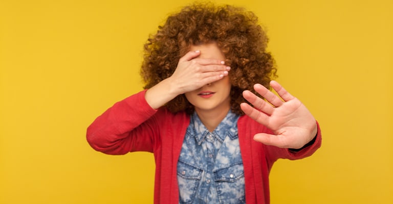 Effective Trade Show Features that Boost Booth Traffic 6 A person with curly hair stands against a bright yellow background, covering their eyes with one hand and holding the other hand out in a gesture that suggests avoidance or reluctance. Their expression and body language convey discomfort or a desire not to see something.