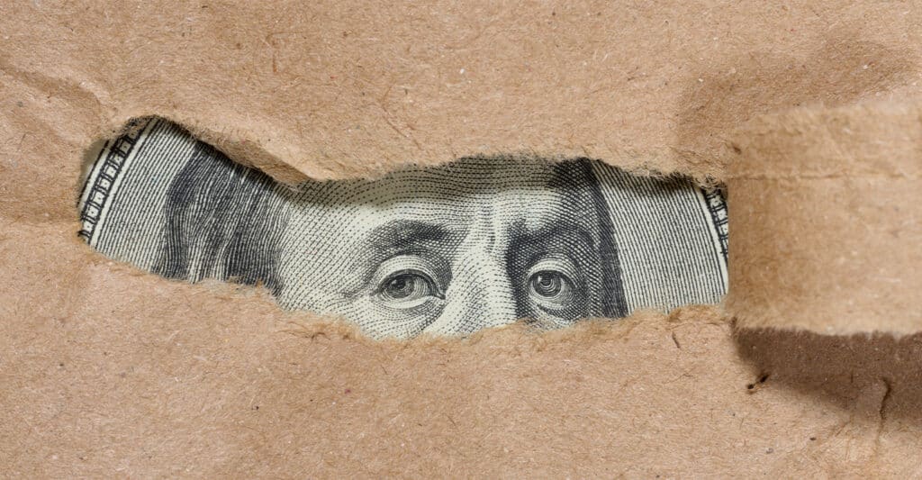 Trade Show Costs: A Complete Breakdown for Exhibitors 4 Close up of torn brown paper revealing the eyes of Benjamin Franklin from a US one hundred dollar bill underneath. The ripped opening focuses on the detailed engraved eyes, creating a concept image about hidden money, secrecy, or financial transparency.