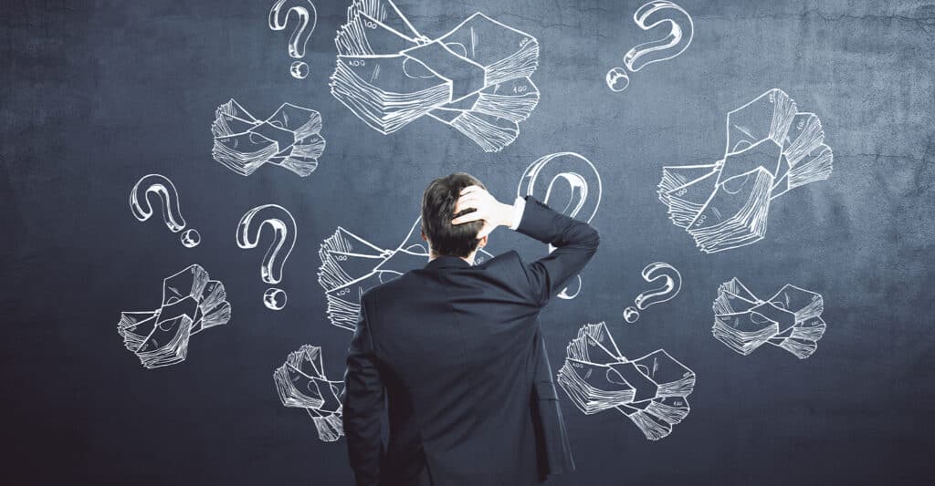 Trade Show Costs: A Complete Breakdown for Exhibitors 5 Man in a business suit stands with his back to the camera, scratching his head while facing a chalkboard covered with drawings of cash bundles and question marks. The chalk illustrations of money and uncertainty symbols create a visual concept of financial confusion, budgeting decisions, or money management challenges.