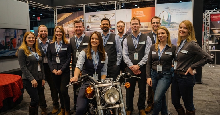 Epic Trade Show Staff: How to Boost Your Booth to the Max 3 A friendly trade show team posing together in front of a branded booth, featuring a motorcycle display.