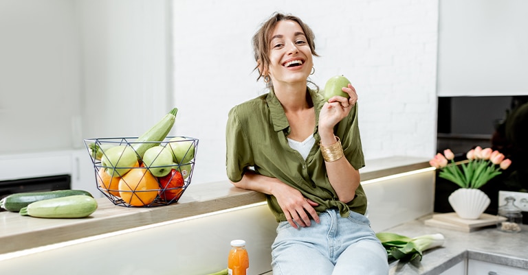 Surefire Trade Show Themes that Will Draw a Big Crowd 7 Smiling woman sitting in a kitchen holding a green apple, with a basket of fresh fruits and vegetables nearby.