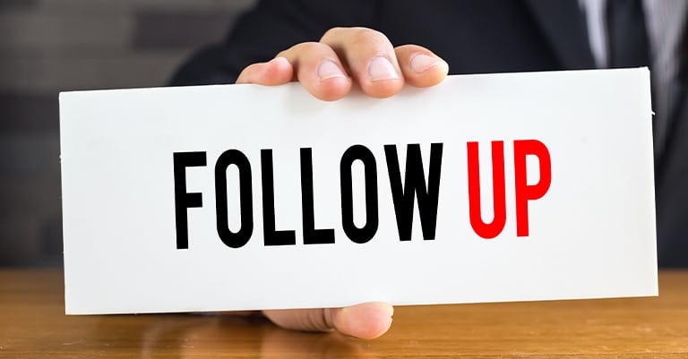 Trade Show Booth Tips: The Ultimate List for Success 8 A hand holding a white sign with the words "Follow Up" in bold black and red text, emphasizing the importance of post-event actions.